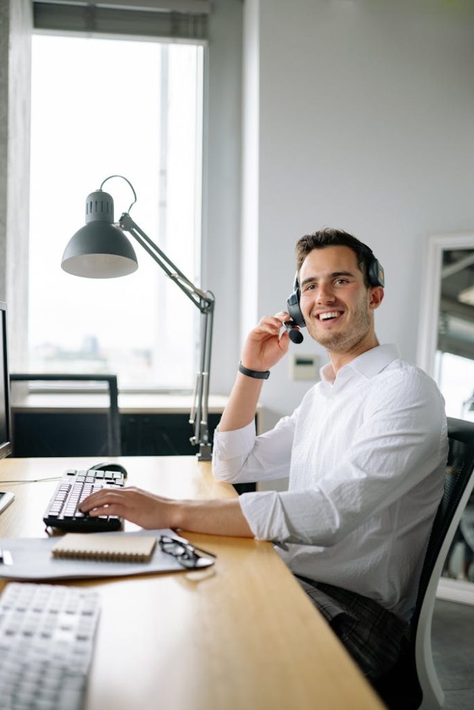 A cheerful call center agent wearing headphones works at a modern office desk.